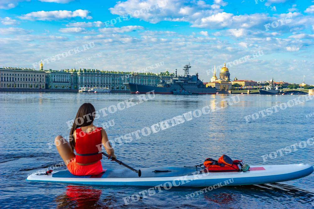 На фотографии изображена женщина, гребущая на SUP-доске по реке Неве с видом на знаменитый исторический центр Санкт-Петербурга. Вдалеке можно увидеть Зимний дворец и шпиль Исаакиевского собора, что придаёт сцене характерный петербургский колорит. Этот снимок может быть использован для статей о городских активностях, в блогах о спорте и туризме, в рекламе спортивного оборудования или как иллюстрация к путеводителям по городу.